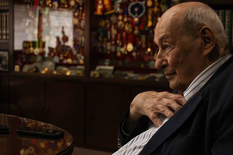 A imagem mostra um homem idoso sentado à mesa, com uma expressão pensativa. Ele está vestido com um paletó escuro e uma camisa listrada. Ao fundo, há uma estante repleta de medalhas e prêmios, refletindo um ambiente de conquistas. O foco da imagem está no homem, enquanto o fundo é desfocado.