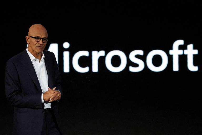 Um homem em um terno escuro está em pé ao lado de um grande logotipo da Microsoft， que está escrito em letras brancas sobre um fundo preto. O homem parece estar se preparando para fazer uma apresentação.