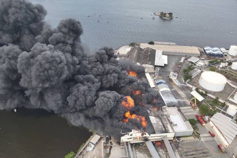 A imagem mostra uma grande nuvem de fumaça preta saindo de um incêndio em uma área industrial. Chamas laranjas são visíveis entre os edifícios， que parecem ser armazéns ou fábricas. Ao fundo， há um corpo d039;água e algumas estruturas industriais， como tanques e barcos.