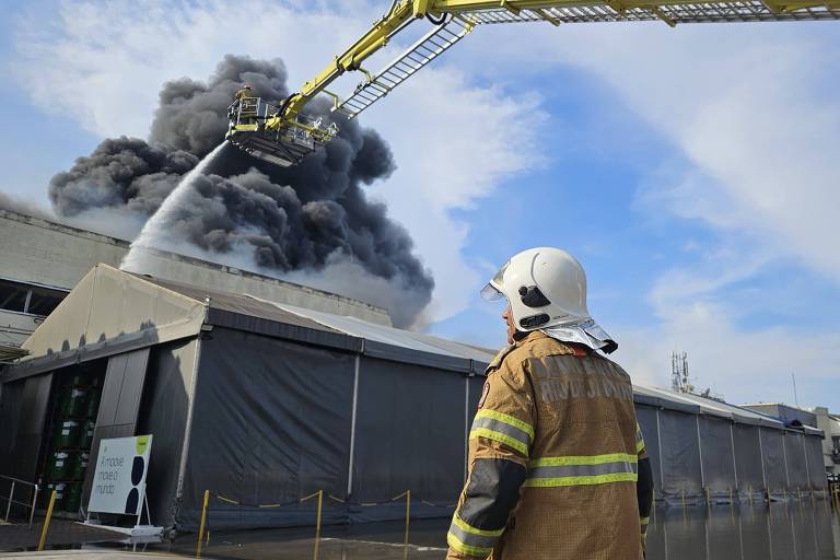 Imagem mostra um bombeiro em primeiro plano， usando um capacete e um uniforme de combate a incêndios. Ele está de costas， observando uma grande nuvem de fumaça preta que se eleva de um edifício em chamas ao fundo. Um caminhão de bombeiros com uma mangueira elevada está direcionando água para o fogo. O céu está parcialmente nublado.