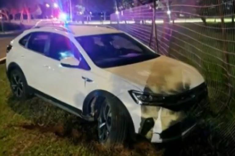 A imagem mostra um carro branco， possivelmente um SUV， que está estacionado em uma área gramada. O veículo apresenta danos visíveis na parte frontal. Ao fundo， há luzes de emergência de um veículo da polícia， indicando que a cena pode estar relacionada a um acidente. É noite， e a iluminação é baixa， com algumas árvores visíveis ao redor.