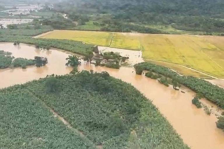 A  imagem mostra uma área rural afetada por inundações， com água marrom cobrindo partes do solo e vegetação. É possível ver campos de cultivo， com uma área verde e outra amarela， além de árvores e uma paisagem montanhosa ao fundo.