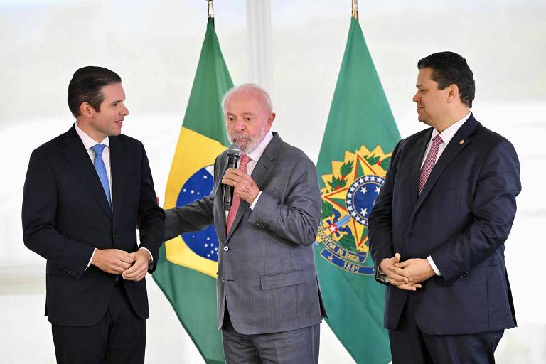 Três homens estão em pé em um ambiente formal. À esquerda， um homem de terno escuro e gravata， à direita， outro homem também de terno escuro. No centro， um homem mais velho， com cabelo grisalho， está falando ao microfone. Atrás deles， estão as bandeiras do Brasil， uma verde e amarela e outra verde com o brasão nacional.