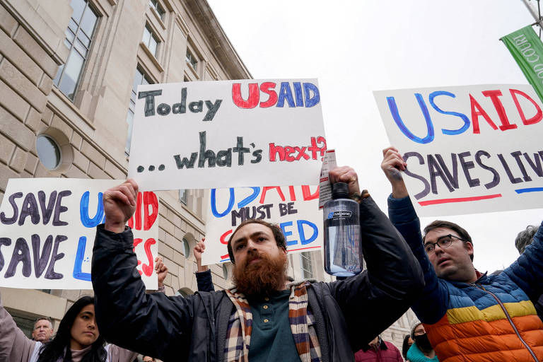 A imagem mostra um grupo de pessoas participando de um protesto. Algumas delas seguram cartazes com mensagens como 039;Today USAID ... what039;s next039; e 039;USAID SAVES LIVES039;. O ambiente parece ser urbano， com edifícios ao fundo. As pessoas estão vestidas de forma casual， e uma delas tem uma barba espessa.