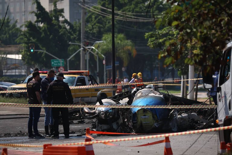 A imagem mostra o local de um acidente aéreo， onde um avião está danificado e parcialmente destruído. Há policiais e equipes de emergência presentes， além de fitas de isolamento ao redor da área. No fundo， é possível ver árvores e sinais de trânsito