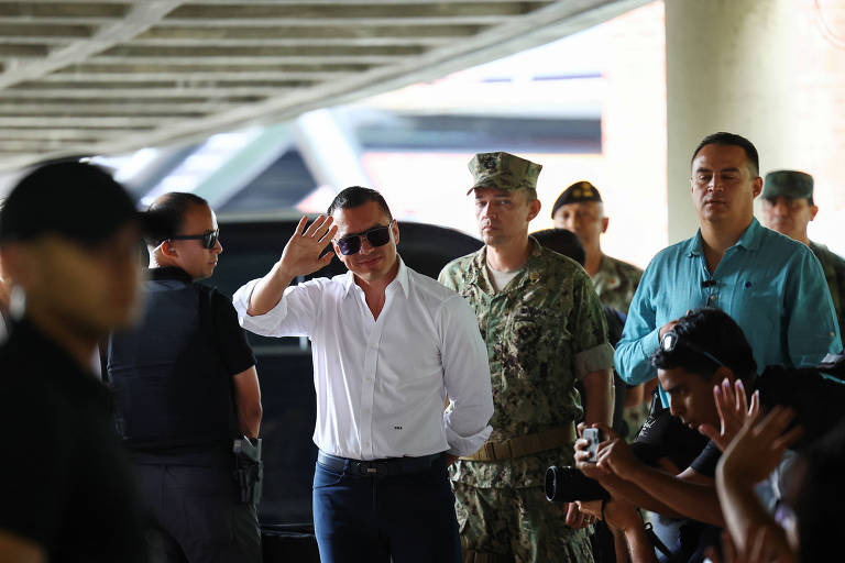 A imagem mostra um grupo de pessoas em um evento público. Um homem de camisa branca e óculos escuros está acenando， enquanto outros homens ao seu redor， incluindo um militar， observam. Há também pessoas na multidão que parecem estar tirando fotos ou acenando. O ambiente é ao ar livre， sob uma estrutura coberta.