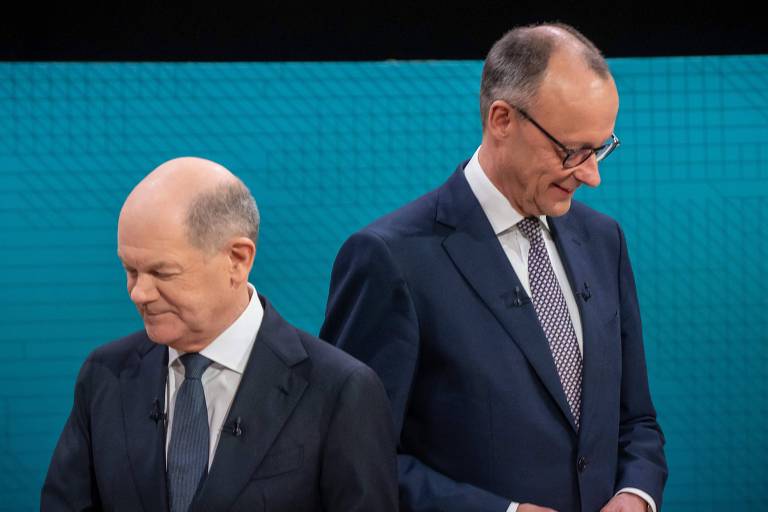 A imagem mostra dois homens em um debate político. O homem à esquerda está com a cabeça baixa， enquanto o homem à direita está ligeiramente inclinado para frente， ambos vestindo ternos escuros. O fundo é de uma cor azul clara.