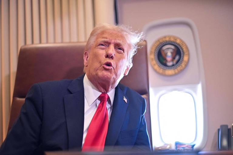 Um homem sentado em uma cadeira dentro de um avião， com cabelo loiro e uma gravata vermelha. Ele parece estar falando ou expressando uma opinião. Ao fundo， há uma parede com cortinas e um emblema que representa a presidência dos Estados Unidos.