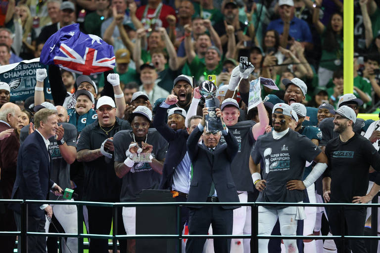 Imagem mostra jogadores de futebol americano， com calças brancas e camisas verdes. São atletas do Philadelphia Eagles， que estão em um palanque comemorando a vitória no campeonato. Ao fundo estão os torcedores. 