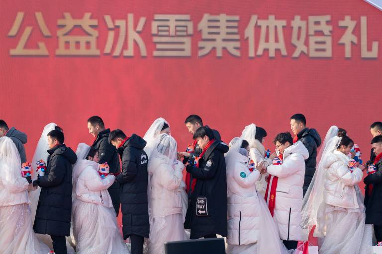 A imagem mostra um grupo de casais em um casamento coletivo， todos vestidos com roupas de inverno. Os noivos usam vestidos brancos e os noivos estão com casacos escuros. Eles estão alinhados em uma cerimônia， segurando pequenos objetos nas mãos. Ao fundo， há uma grande faixa vermelha com caracteres chineses.