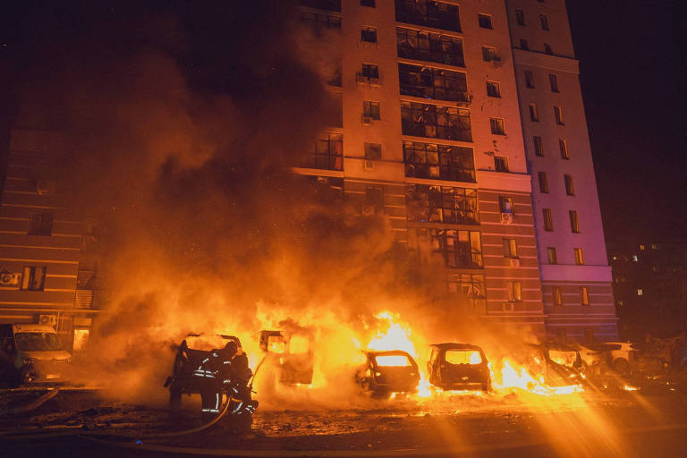 Bombeiros trabalham contra incêndio em carros na frente de um prédio residencial à noite