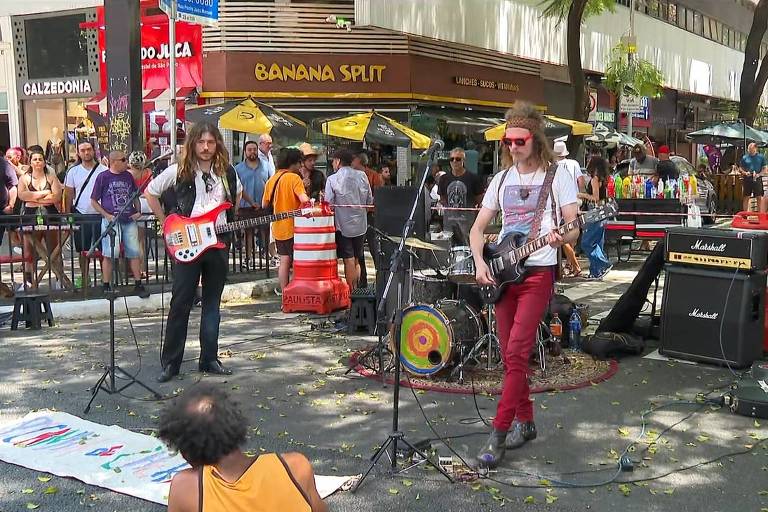 A imagem mostra uma banda de rua se apresentando em um ambiente urbano. Dois músicos estão em destaque， um tocando guitarra e o outro em um microfone， enquanto um terceiro membro está atrás de um kit de bateria. O público está ao fundo， com várias pessoas observando a performance. Há barracas e lojas visíveis ao redor， e a cena é iluminada pela luz do dia.