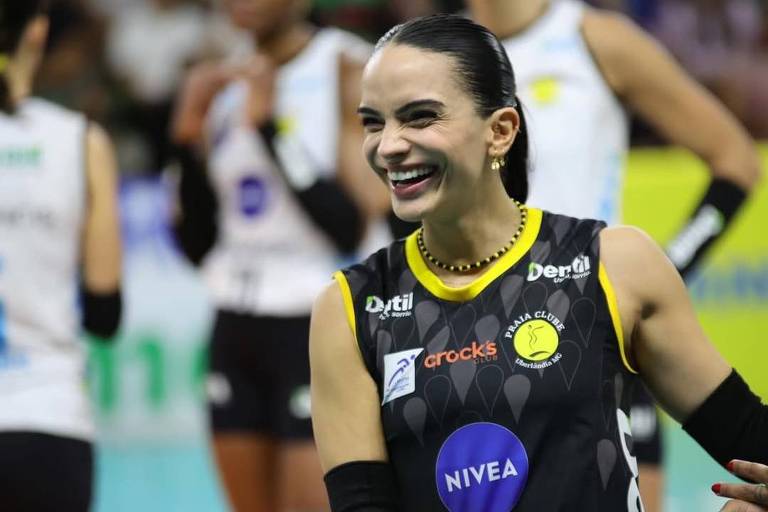 Uma jogadora de vôlei está sorrindo e interagindo com outras jogadoras em um ambiente de competição. Ela usa uma camisa preta com detalhes amarelos e patrocinadores visíveis, como Nivea e Neutrox. Ao fundo, outras jogadoras estão em uniformes brancos, e a atmosfera é de celebração e camaradagem.