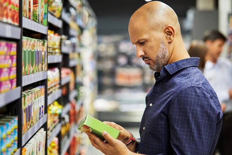 Um homem calvo, vestindo uma camisa azul, está em um supermercado, olhando atentamente para um produto verde que segura em suas mãos. Ao fundo, prateleiras repletas de produtos variados estão visíveis, e há outras pessoas fazendo compras.