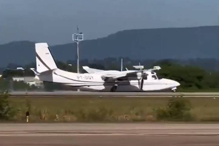 Um avião de asas altas， predominantemente branco， está estacionado em uma pista. Ao fundo， há uma paisagem montanhosa sob um céu claro. A imagem mostra a lateral do avião， que possui detalhes em vermelho nas asas.