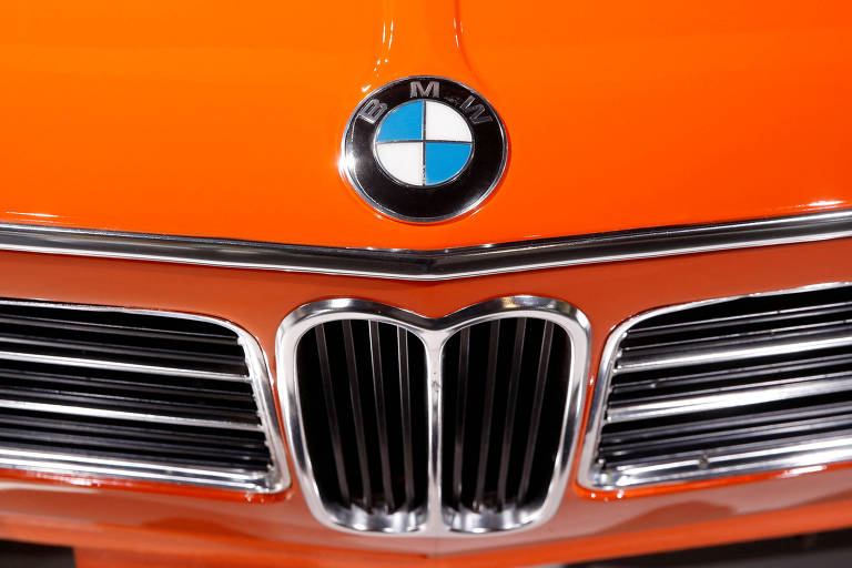 A imagem mostra a frente de um carro BMW， destacando o logotipo da marca em um fundo laranja. O logotipo é circular， com as cores azul e branca， e está posicionado acima da grade frontal， que possui um design característico com barras verticais. A grade é emoldurada por um acabamento cromado.
