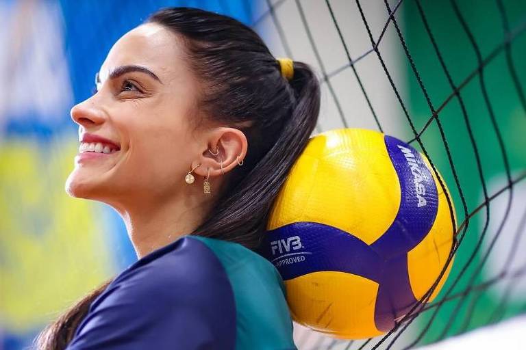 Uma jogadora de vôlei está sorrindo enquanto apoia a cabeça em uma bola de vôlei amarela e roxa, que está encostada na rede. Ela usa uma camiseta azul com detalhes verdes e tem o cabelo preso em um rabo de cavalo. O fundo apresenta uma parede com cores vibrantes, incluindo azul e amarelo, com a palavra 'Brasil' visível.