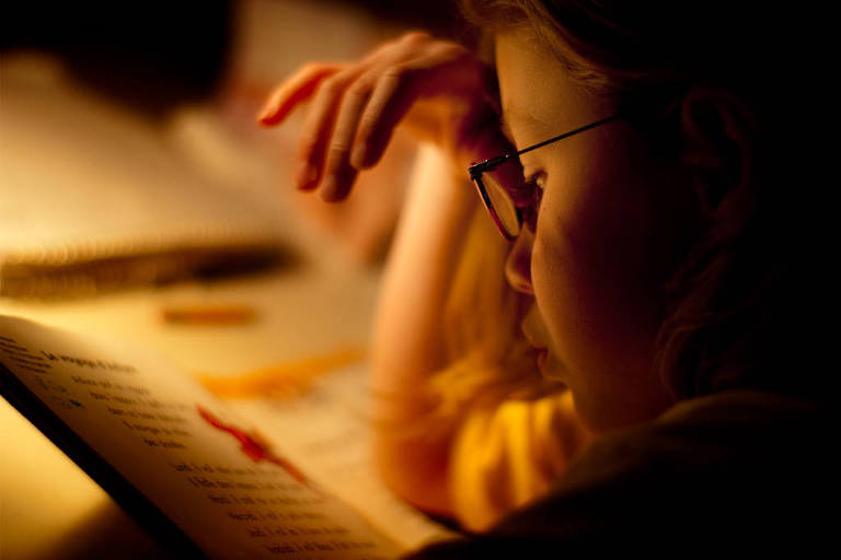 A imagem mostra uma criança concentrada em um livro， com uma mão apoiada na cabeça e a outra segurando uma caneta. A iluminação é suave， destacando o rosto da criança e o livro aberto à sua frente. O fundo é desfocado， sugerindo um ambiente de estudo tranquilo.
