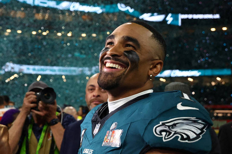 Um jogador de futebol americano， vestindo um uniforme verde com o logotipo dos Philadelphia Eagles， sorri amplamente enquanto celebra. Ele tem pintura facial preta e está cercado por pessoas， incluindo um fotógrafo. O fundo mostra uma atmosfera festiva com confetes caindo e luzes brilhantes.