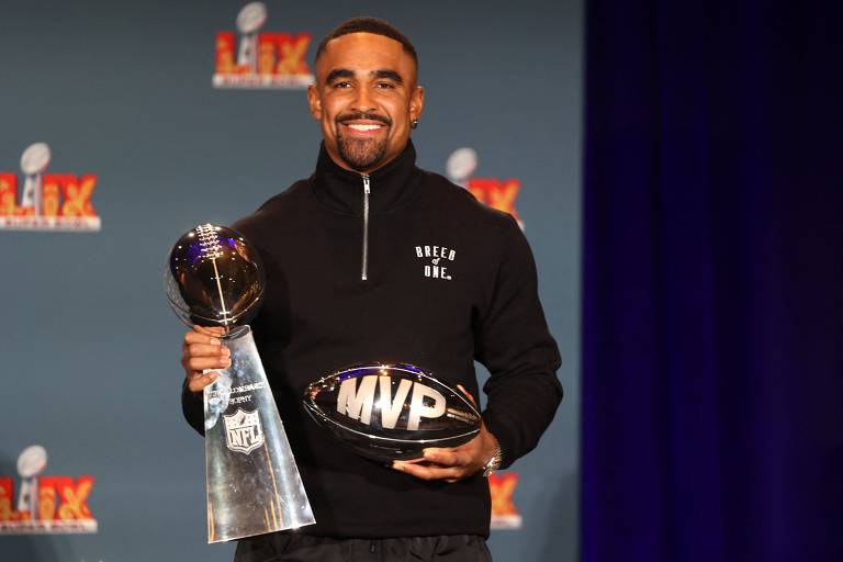 Um jogador de futebol americano está posando para a foto， segurando um troféu da NFL e uma bola com a inscrição 039;MVP039;. Ele está vestindo um suéter preto com a frase 039;Breed of One039;. O fundo é azul com logotipos da NFL e do evento.