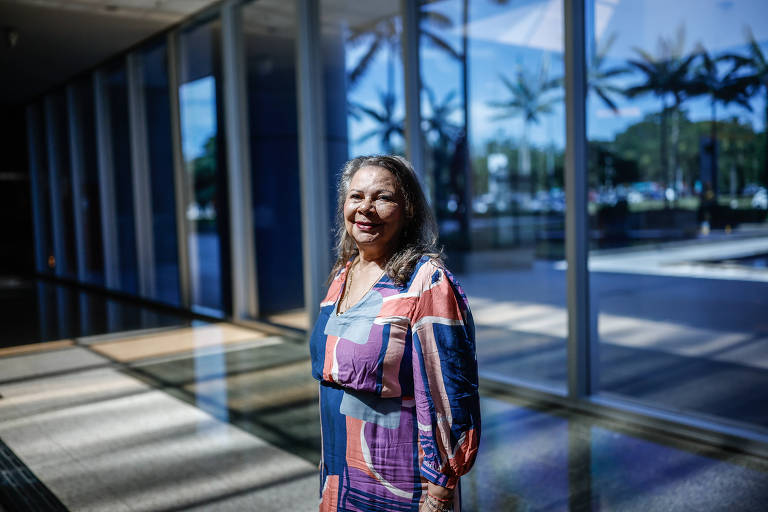 A imagem mostra uma mulher sorrindo em um ambiente com grandes janelas que permitem a entrada de luz natural. Ela está vestindo uma blusa colorida com padrões variados. Ao fundo， há uma vista de palmeiras e um espaço aberto.
