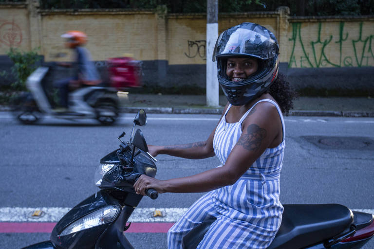 Uma mulher está pilotando uma motocicleta preta， usando um capacete. Ela está sorrindo e vestindo um macacão listrado. Ao fundo， há uma outra motocicleta e uma parede com grafite. A cena parece ser em uma rua urbana.