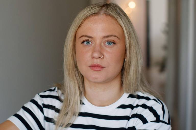 A imagem mostra uma mulher com cabelo loiro e liso， usando uma blusa branca com listras pretas. Ela está olhando para a câmera com uma expressão séria， em um ambiente interno com paredes claras e um fundo desfocado.
