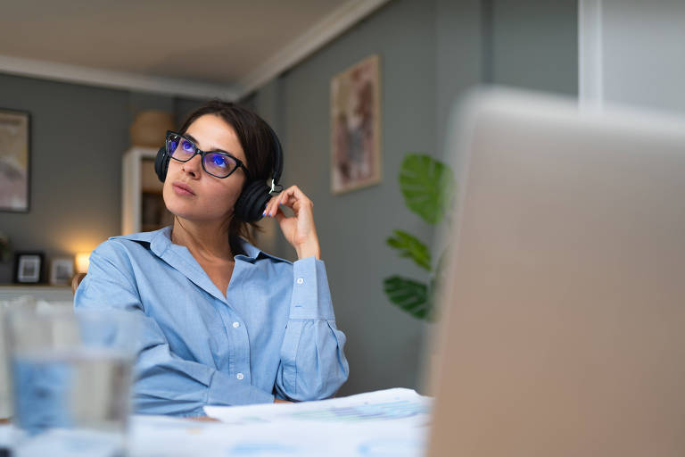 A imagem mostra uma mulher com cabelo escuro e óculos， usando fones de ouvido， olhando para cima com uma expressão pensativa. Ela está vestindo uma camisa azul clara e está sentada em uma mesa com papéis e um copo de água. Ao fundo， há uma planta e algumas decorações na parede.