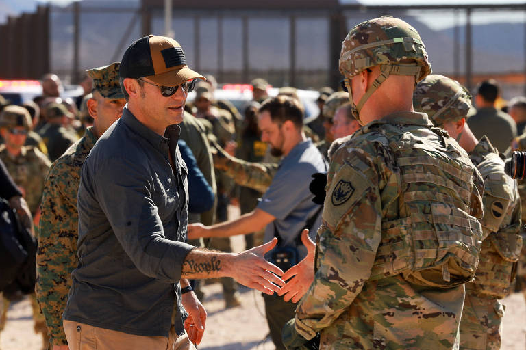 A imagem mostra um grupo de pessoas em um ambiente ao ar livre， onde um homem de camiseta escura e boné está cumprimentando um soldado vestido com uniforme militar. Ao fundo， há outras pessoas e uma cerca， sugerindo um contexto de segurança ou militar. A cena parece ser de um evento ou visita oficial