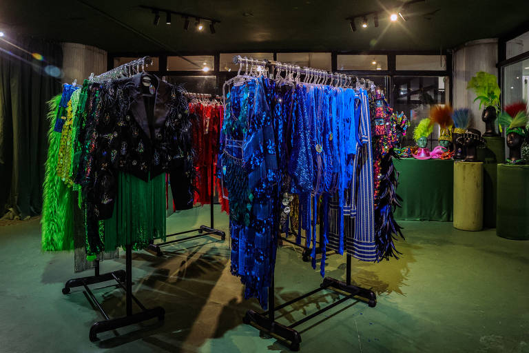 A imagem mostra um espaço de exposição com várias roupas penduradas em cabides. Há roupas em cores vibrantes, como verde, vermelho e azul, organizadas em suportes. Ao fundo, há uma mesa com objetos decorativos coloridos, incluindo itens em verde e rosa. A iluminação é suave, destacando as roupas.