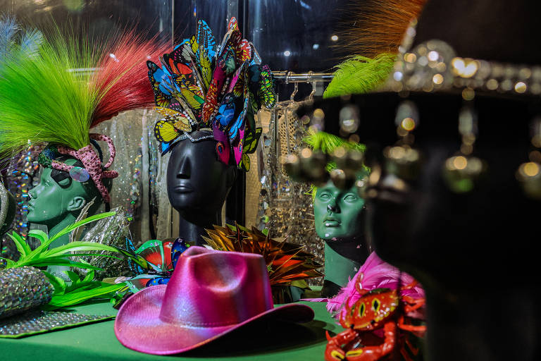 A imagem mostra uma coleção vibrante de acessórios de Carnaval. À esquerda， há máscaras decorativas com penas coloridas e designs intricados， evocando um visual exótico e festivo. Ao centro， um chapéu rosa brilhante adiciona um toque de cor suave em contraste com os tons mais escuros das máscaras. À direita， outros adereços carnavalescos， como leques e mais máscaras， são visíveis， todos ricamente adornados e prontos para animar qualquer celebração de Carnaval. O fundo escuro realça as cores vivas e os detalhes metálicos dos acessórios， criando uma atmosfera festiva e alegre.








