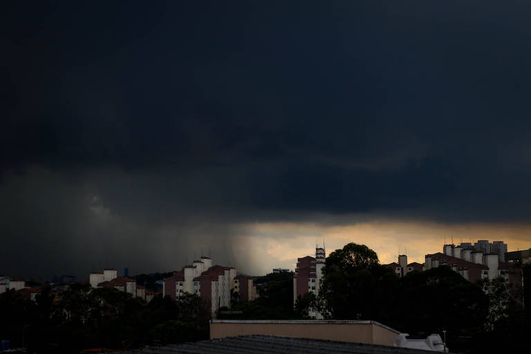 A imagem mostra um céu escuro e nublado， indicando a aproximação de uma tempestade. No horizonte， há uma faixa de luz que sugere a presença do sol， enquanto a chuva parece cair em uma área distante. A silhueta de prédios urbanos é visível na parte inferior da imagem， contrastando com o céu ameaçador.