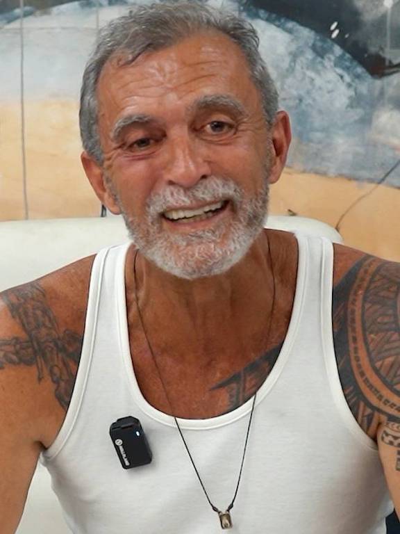 Um homem de pele clara e cabelo grisalho, com barba e tatuagens visíveis nos braços, está sorrindo. Ele usa uma camiseta branca sem mangas e um colar. Um pequeno dispositivo preto está preso à sua camiseta. Ao fundo, há uma parede com uma pintura abstrata