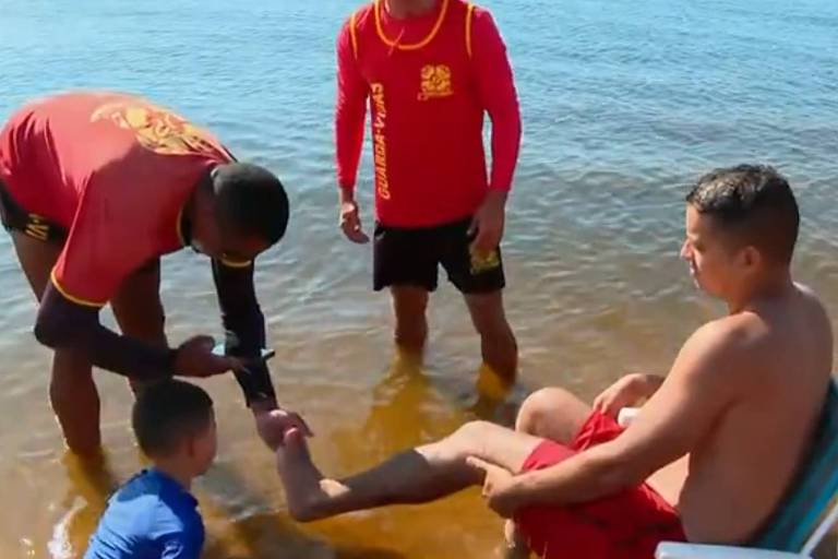 A imagem mostra um grupo de quatro pessoas em uma praia. Dois homens， vestidos com camisetas vermelhas de salva-vidas， estão ajudando um homem sentado em uma cadeira de praia. O homem sentado， que está sem camisa e usando shorts vermelhos， parece estar recebendo assistência em seu pé. Um menino， vestido com uma camiseta azul， está próximo， observando a situação. O ambiente é de água rasa e clara， com um céu azul ao fundo.