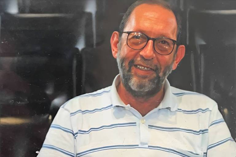 Um homem de meia-idade com barba e óculos， sorrindo. Ele está usando uma camisa listrada em branco e azul. O fundo é desfocado， com cadeiras escuras visíveis.