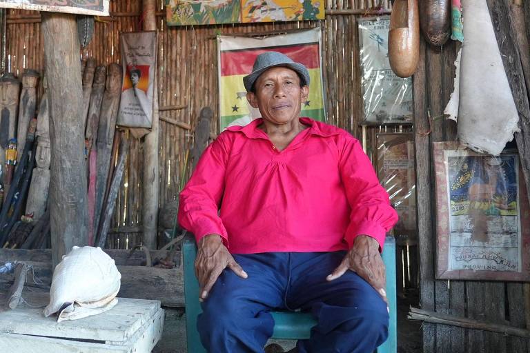 Um homem sentado em uma cadeira em um ambiente rústico. Ele usa uma camisa rosa e um chapéu. Ao fundo， há várias ferramentas penduradas e cartazes nas paredes. Na mesa à sua frente， há um objeto que parece ser um capacete e outros itens não identificáveis.