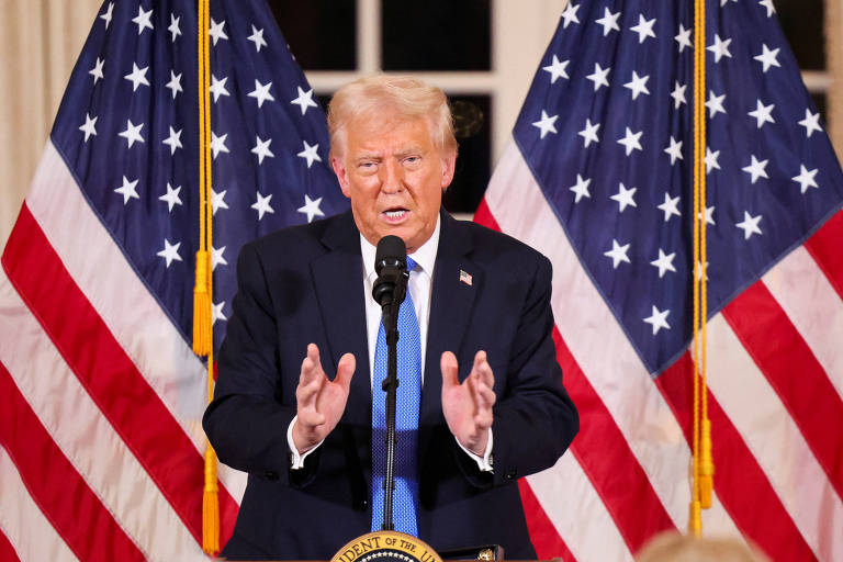 A imagem mostra Donald Trump falando em um evento oficial. Ele está em frente a duas grandes bandeiras dos Estados Unidos. Trump está usando um terno escuro， uma camisa branca e uma gravata azul. Ele parece estar fazendo um discurso， com as mãos levantadas em um gesto de ênfase. O cenário sugere um ambiente formal， possivelmente um evento político ou uma coletiva de imprensa.