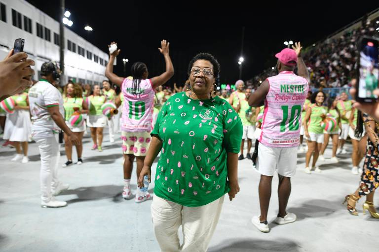 A imagem mostra uma mulher sorridente vestindo uma camisa verde com estampas， caminhando em um ambiente de festa. Ao fundo， há várias pessoas dançando， vestindo roupas coloridas， incluindo camisetas rosa e verde. O cenário é iluminado por luzes artificiais， sugerindo que a cena ocorre à noite durante um evento festivo.