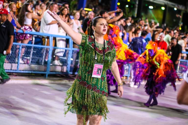 Uma mulher vestida com um traje verde de penas dança em um desfile de carnaval. Ela está levantando um braço e parece estar se divertindo. Ao fundo， há outras pessoas dançando， algumas vestindo trajes coloridos e vibrantes， com plumas e adereços. O ambiente é festivo， com uma multidão assistindo ao desfile.