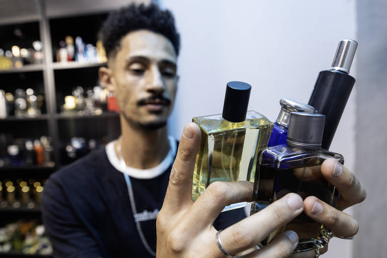 Um homem está segurando quatro frascos de perfume em suas mãos. Ele está em um ambiente com prateleiras ao fundo， repletas de outros frascos de perfume. O homem tem cabelo crespo e usa uma camiseta escura. Ele parece estar avaliando os frascos que está segurando.
