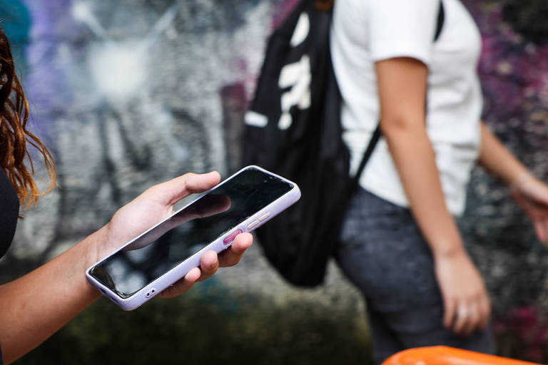 A imagem mostra uma mão segurando um celular com uma capa roxa. Ao fundo， uma pessoa de camiseta branca e mochila preta está caminhando， enquanto uma parede grafitada é visível atrás delas.