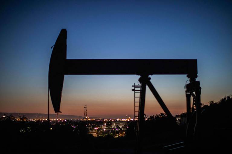 A imagem mostra uma bomba de petróleo em silhueta contra um céu ao entardecer， com tons de azul e laranja. A estrutura da bomba é visível， assim como uma escada ao lado. Ao fundo， há uma cidade iluminada