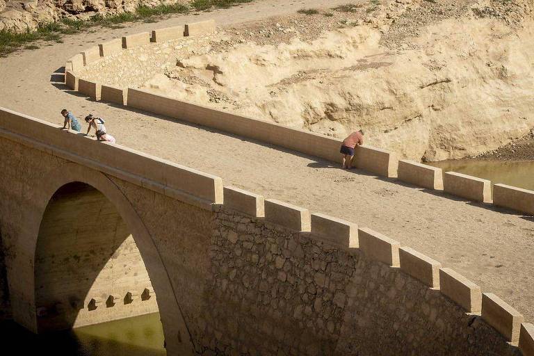 A imagem mostra uma ponte de pedra que se curva sobre um rio. Algumas pessoas estão caminhando sobre a ponte， enquanto outras estão sentadas em um dos lados. O cenário ao fundo apresenta uma encosta de rochas e vegetação escassa， com um tom de terra clara. A água do rio é visível embaixo da ponte.