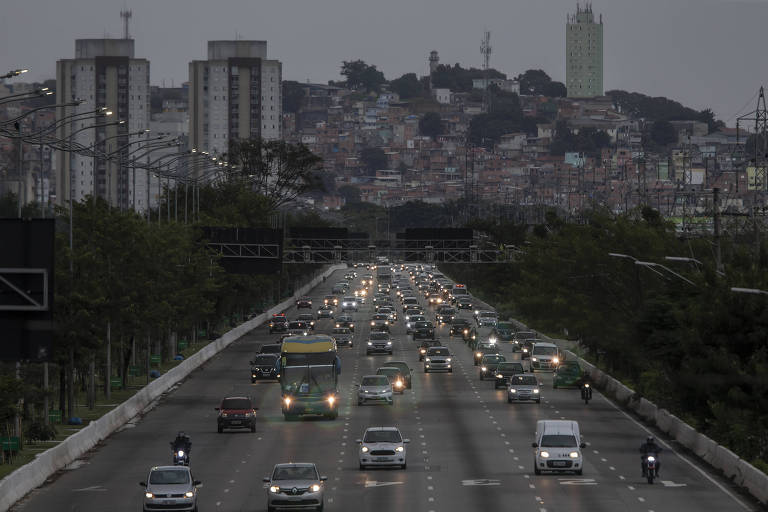 A imagem mostra uma rodovia com tráfego intenso em um dia nublado. Vários veículos estão se deslocando em ambas as direções， e ao fundo， é possível ver edifícios e uma colina com casas. O céu está escuro， sugerindo as primeiras horas do dia