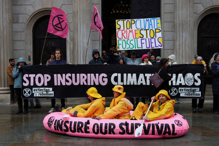 A imagem mostra um protesto em frente a um edifício. No centro， três pessoas vestidas com trajes amarelos estão sentadas em um bote inflável rosa. Atrás delas， um grande banner preto diz 039;STOP INSURING CLIMATE CHAOS039;. Algumas pessoas seguram cartazes， incluindo um que diz 039;STOP CLIMATE CRIMES STOP FOSSIL FUELS039;. O ambiente parece chuvoso， com várias pessoas usando capas de chuva.