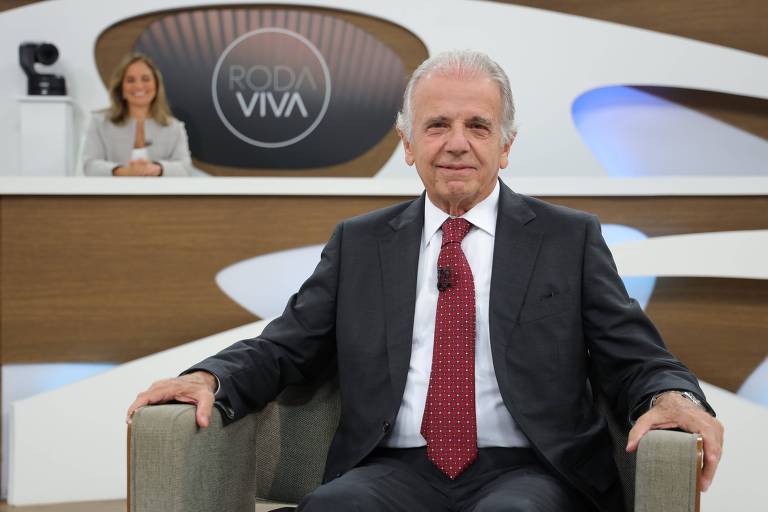 Um homem de terno escuro e gravata vermelha está sentado em um sofá, olhando para a câmera. Ao fundo, uma mulher está em uma mesa, visível parcialmente, com um logotipo que diz 'VIVA' em um painel circular. O ambiente é moderno e bem iluminado.