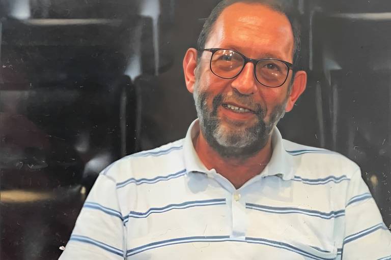 Um homem de meia-idade com barba e óculos, sorrindo. Ele está usando uma camisa listrada em branco e azul. O fundo é desfocado, com cadeiras escuras visíveis.