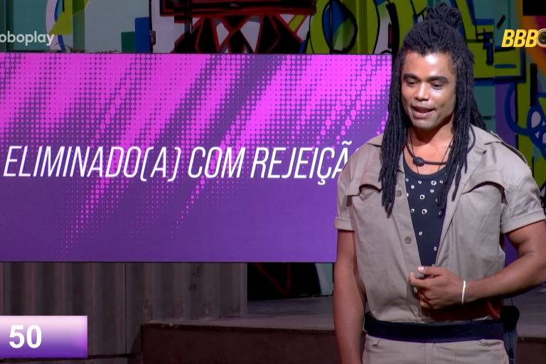 Um homem com cabelo longo e dreadlocks está em pé, olhando para a câmera. Ele usa uma camisa de manga curta bege e uma camiseta preta com detalhes. Ao fundo, uma tela grande exibe a frase 'ELIMINADO(A) COM REJEIÇÃO' em letras brancas sobre um fundo roxo. Na parte inferior da tela, há um número '50' em um fundo lilás.