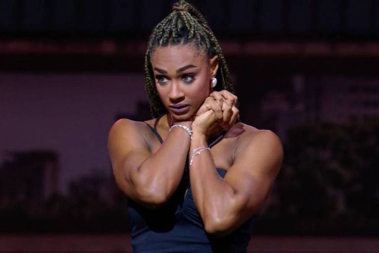 A imagem mostra uma mulher com cabelo trançado, usando uma blusa preta. Ela está em uma pose expressiva, com os braços cruzados sobre o peito e uma expressão facial que transmite emoção. O fundo é desfocado, sugerindo um ambiente de apresentação ao vivo.