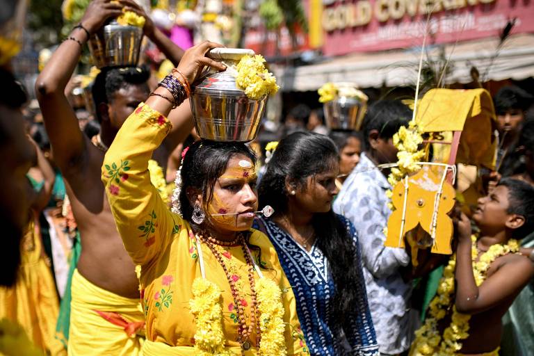 Grupo de mulheres com roupas típicas da cultura hindu realizam uma procissão, e algumas delas carregam uma urna metálica sobre a cabeça